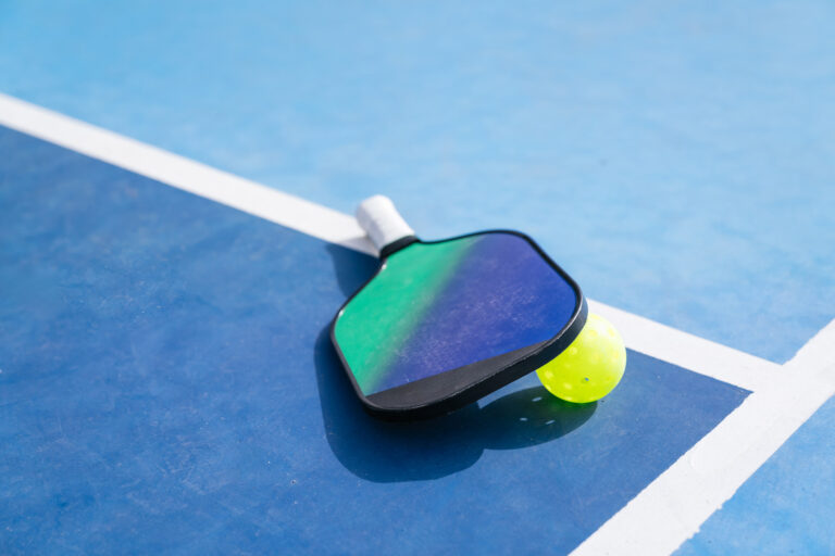 Pickleball paddle and ball resting on a blue court, close-up view near the sideline