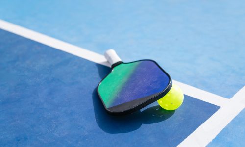 A close-up of a pickleball paddle and ball resting on a blue court near the sideline. Concept of sports equipment in action.