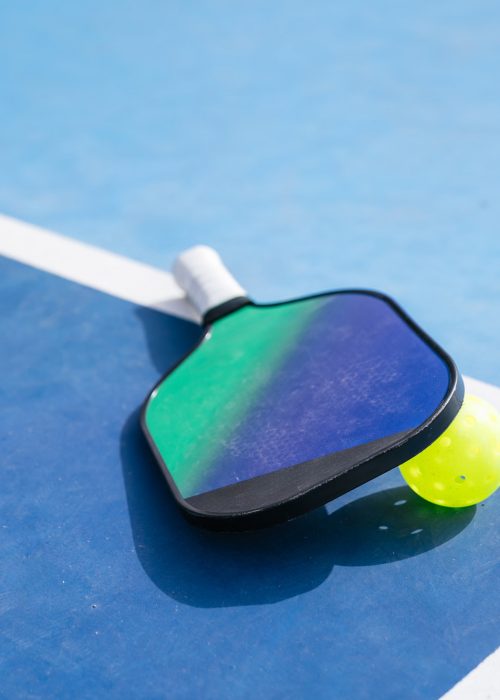 A close-up of a pickleball paddle and ball resting on a blue court near the sideline. Concept of sports equipment in action.